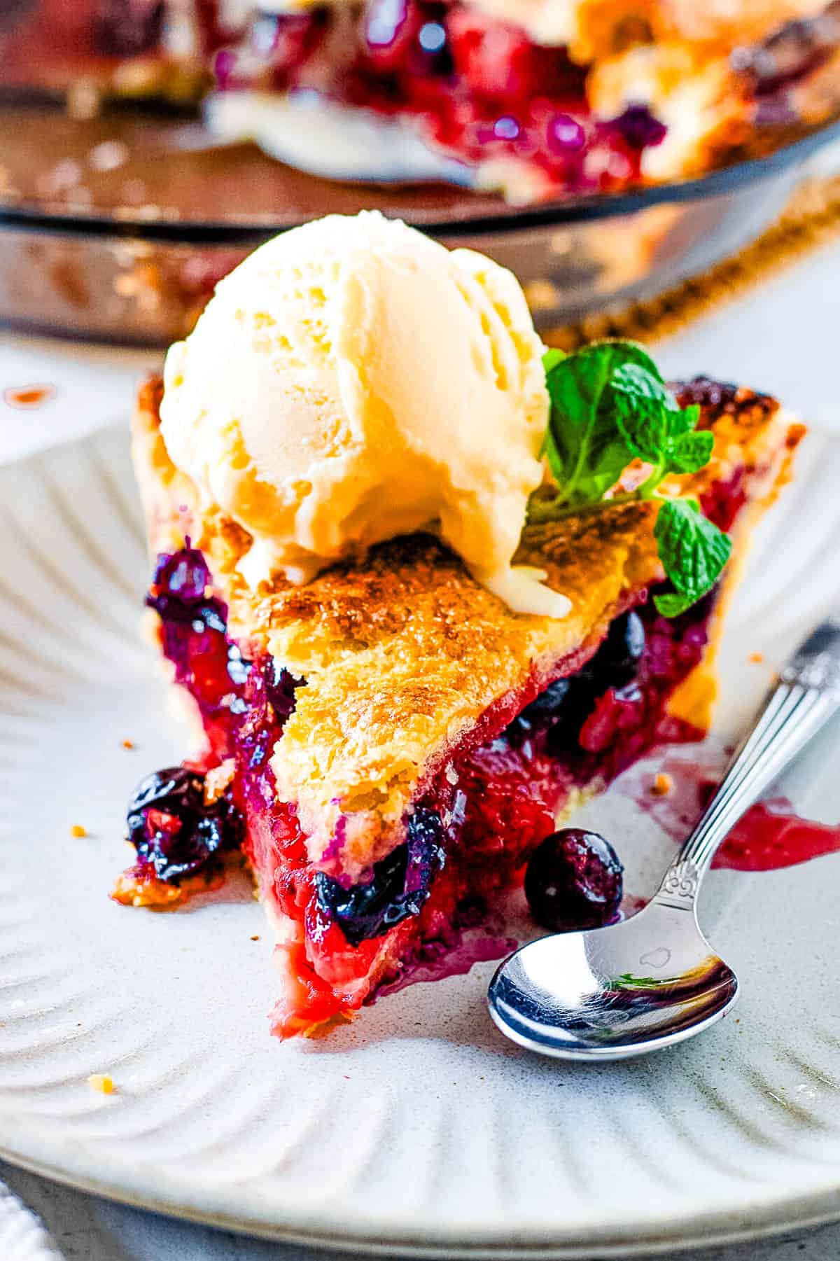 Slice of easy strawberry blueberry pie topped with ice cream with a w،le pie in the background.