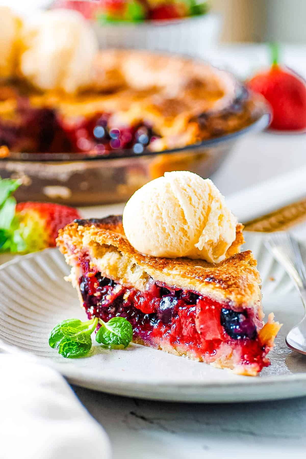 Slice of easy strawberry blueberry pie topped with ice cream with a w،le pie in the background.