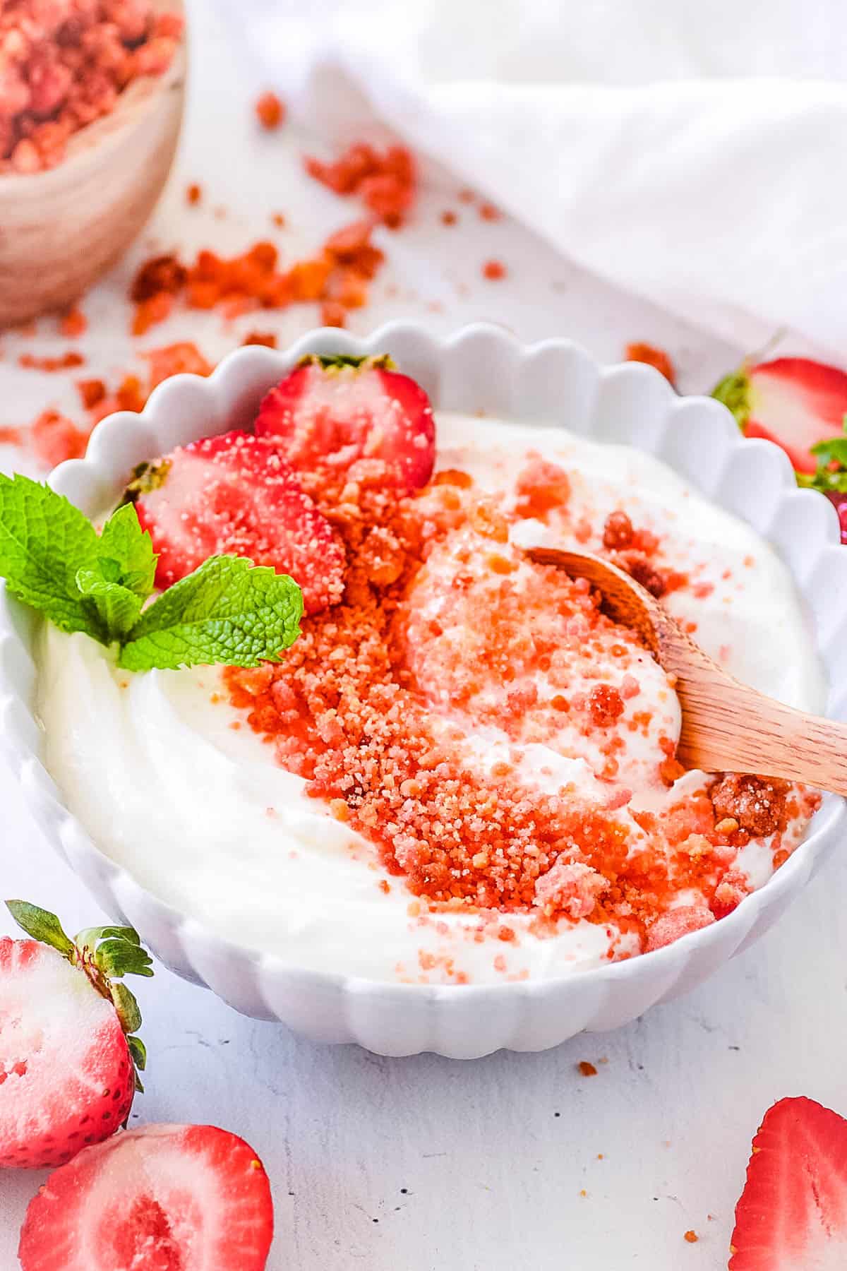 Strawberry crumble topping added on top of yogurt, in a white bowl, garnished with strawberries and mint.