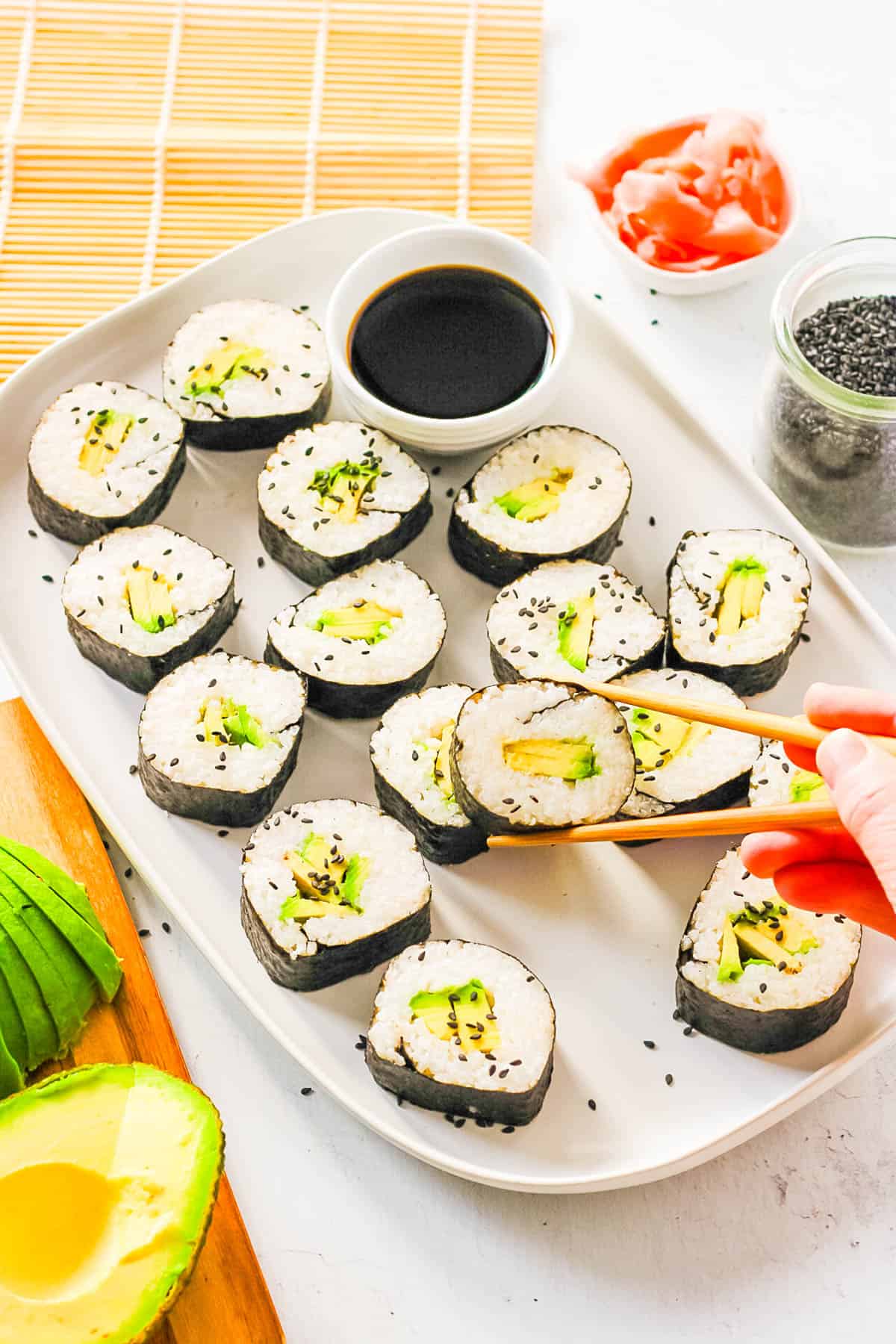 A hand using chopsticks to hold one of the avocado sushi pieces over a platter filled with sliced avocado sushi.