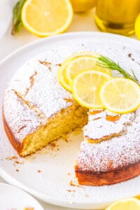 Lemon vegan olive oil cake on a cake stand, garnished with lemon slices and herbs.