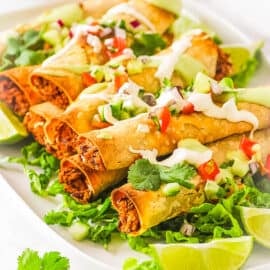 Easy vegan taquitos topped with vegan sour cream, salsa and served on a white platter with lettuce and limes.