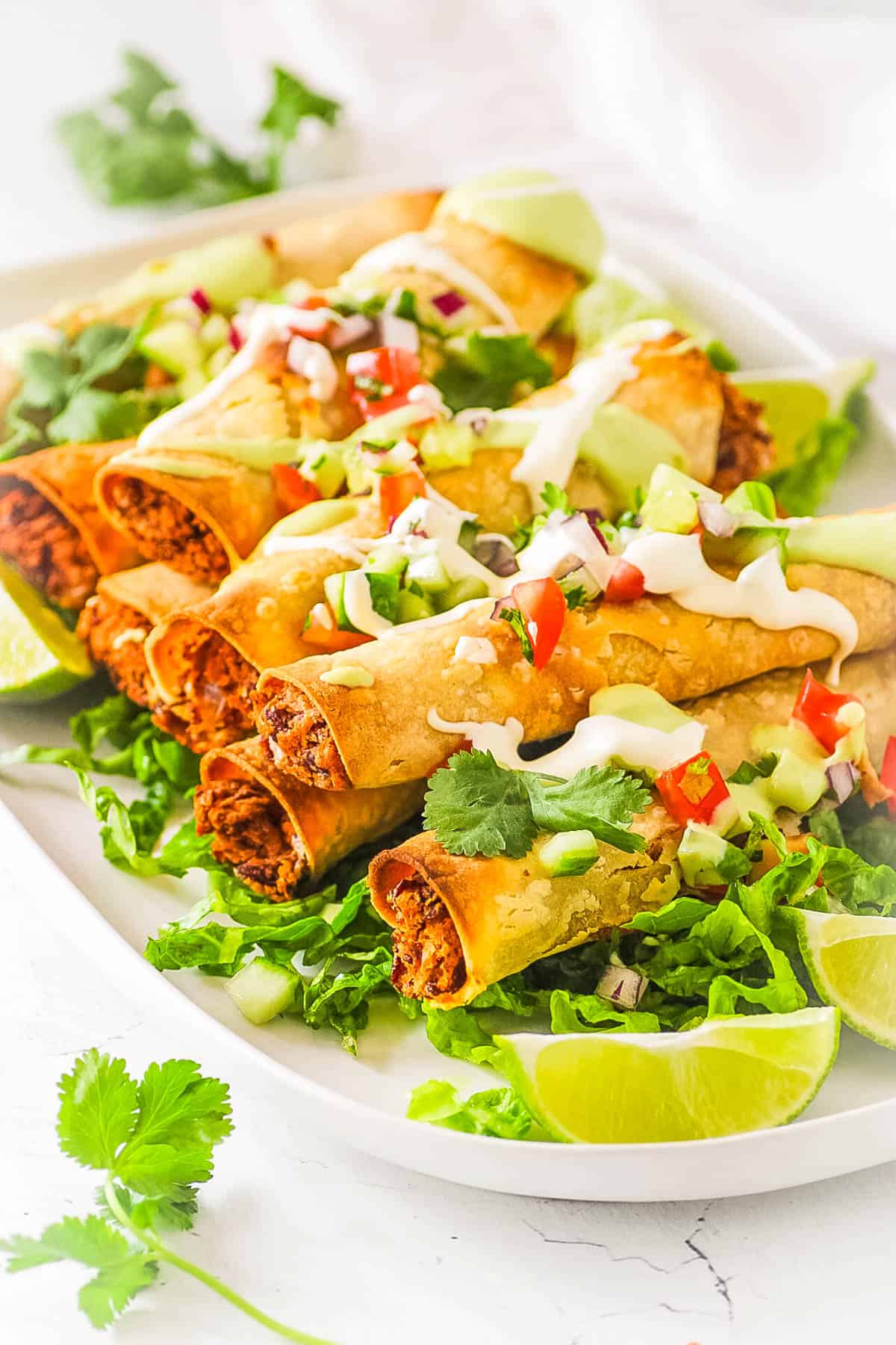 Easy vegan taquitos topped with vegan sour cream, salsa and served on a white platter with lettuce and limes.
