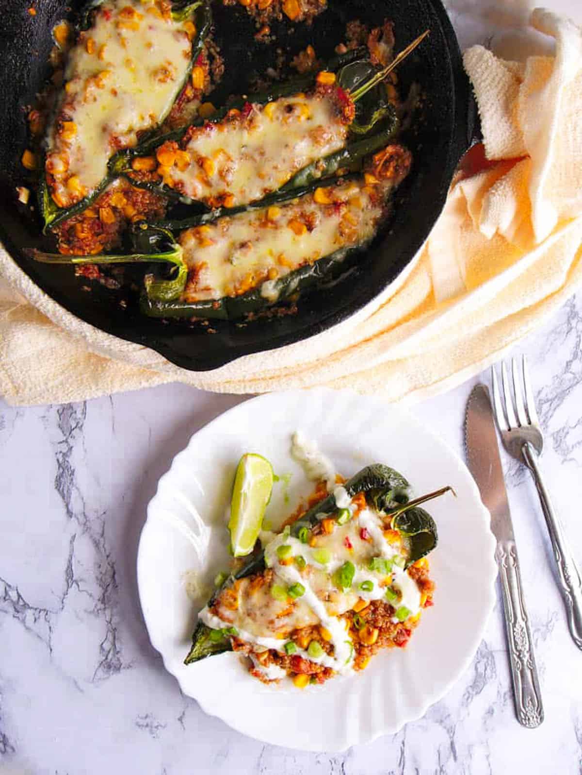 Vegetarian stuffed poblano peppers with melted cheese on top, served in a cast iron skillet with one served on a white plate on the side - drizzled with sour cream, topped with chives, lime wedge on the side - top view