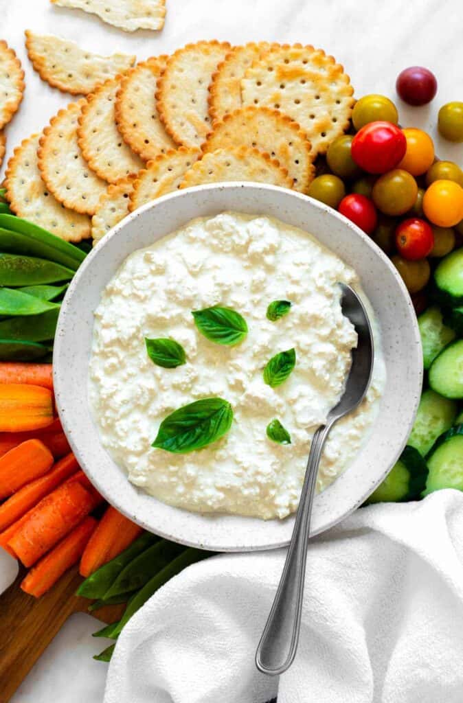Cottage cheese in a white bowl garnished with basil leaves, surrounded by fresh veggies and crackers.