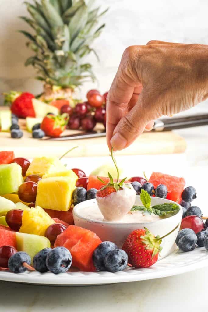 Strawberry being dipped into a vegan fruit dip, surrounded by a variety of fruits.