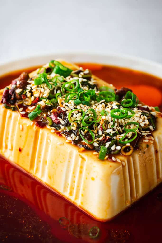 Cold block of silken tofu topped with a soy sauce mixture and ،es, green onion, and sesame seeds.