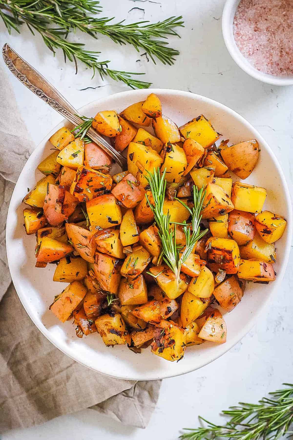 Fried potatoes in a white bowl with a spring of rosemary on top.