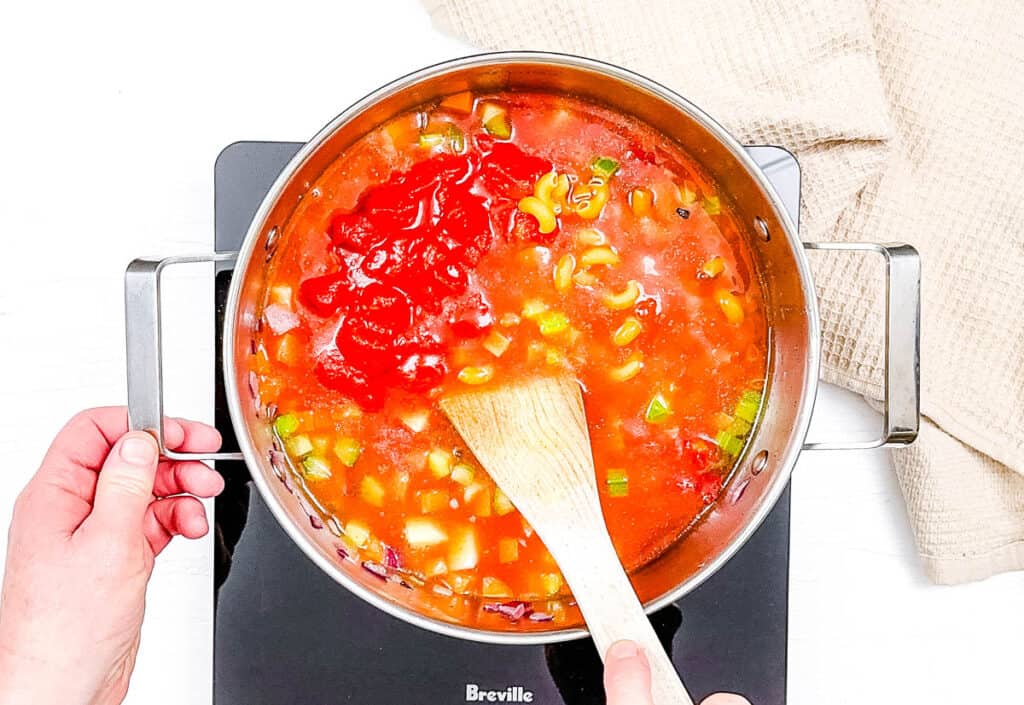 Tomatoes and broth added to veggies and beans cooking in a large stock pot on the stove.