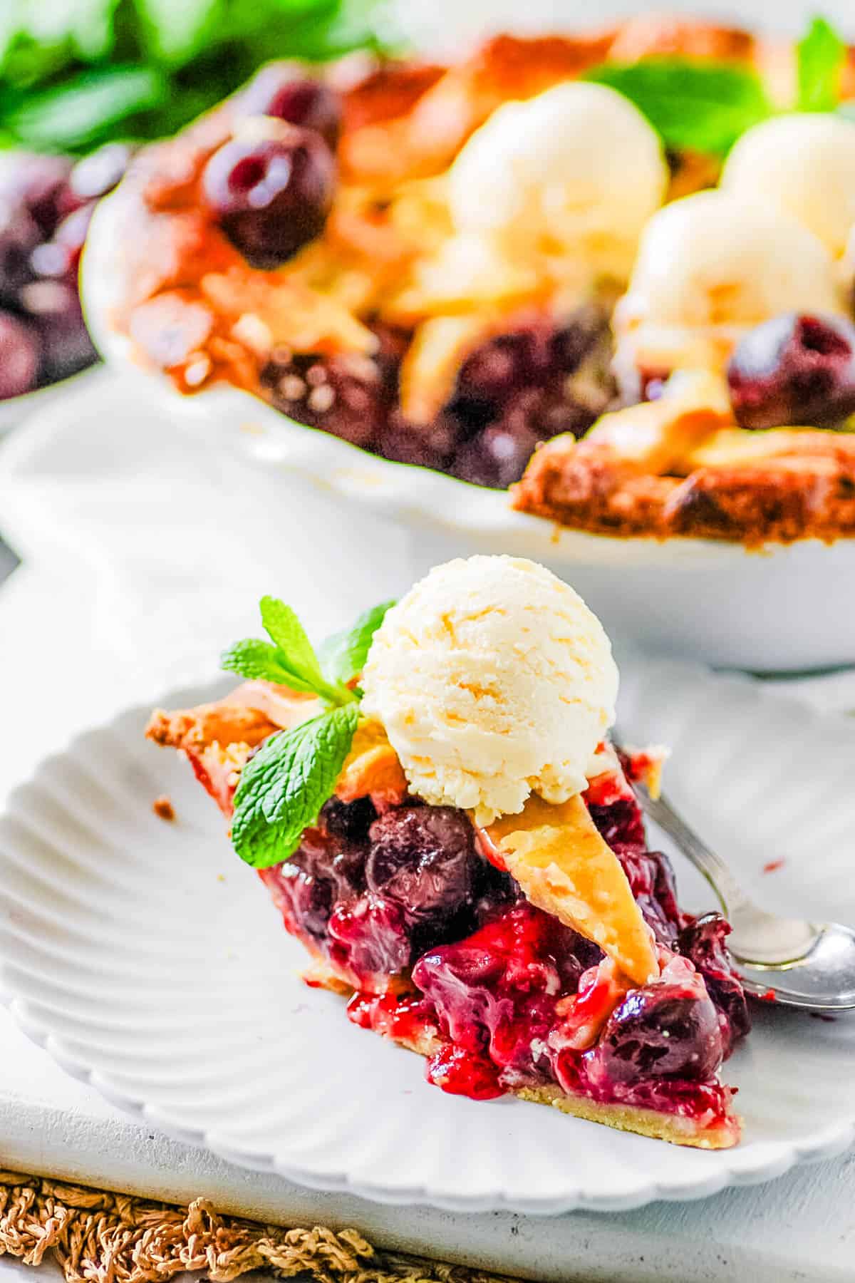 Slice of cherry pie with frozen cherries on a white plate, topped with ice cream and garnished with mint.