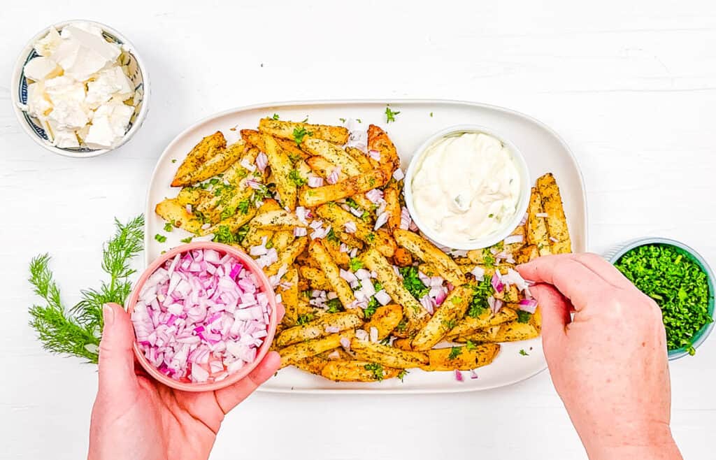 Greek fries with feta cheese, onions, parsley and tzatziki served on a white plate.