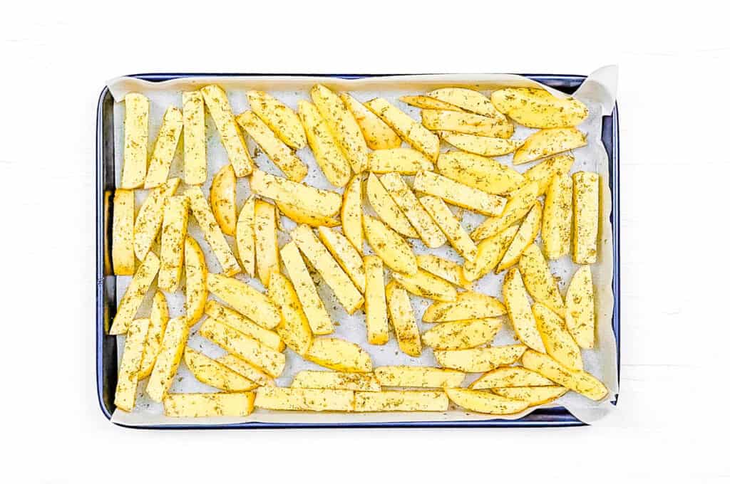 Potato wedges tossed with Greek seasonings and olive oil, spread on a baking sheet lined with parchment paper.