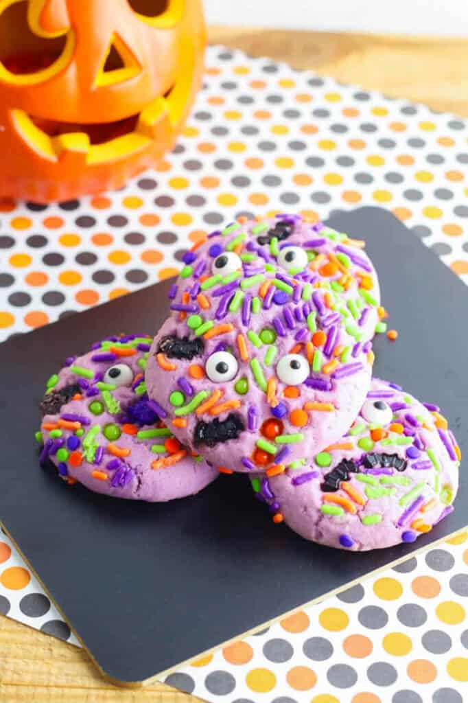 Purple Halloween monster cookies with googley eyes stacked on a black cutting board.
