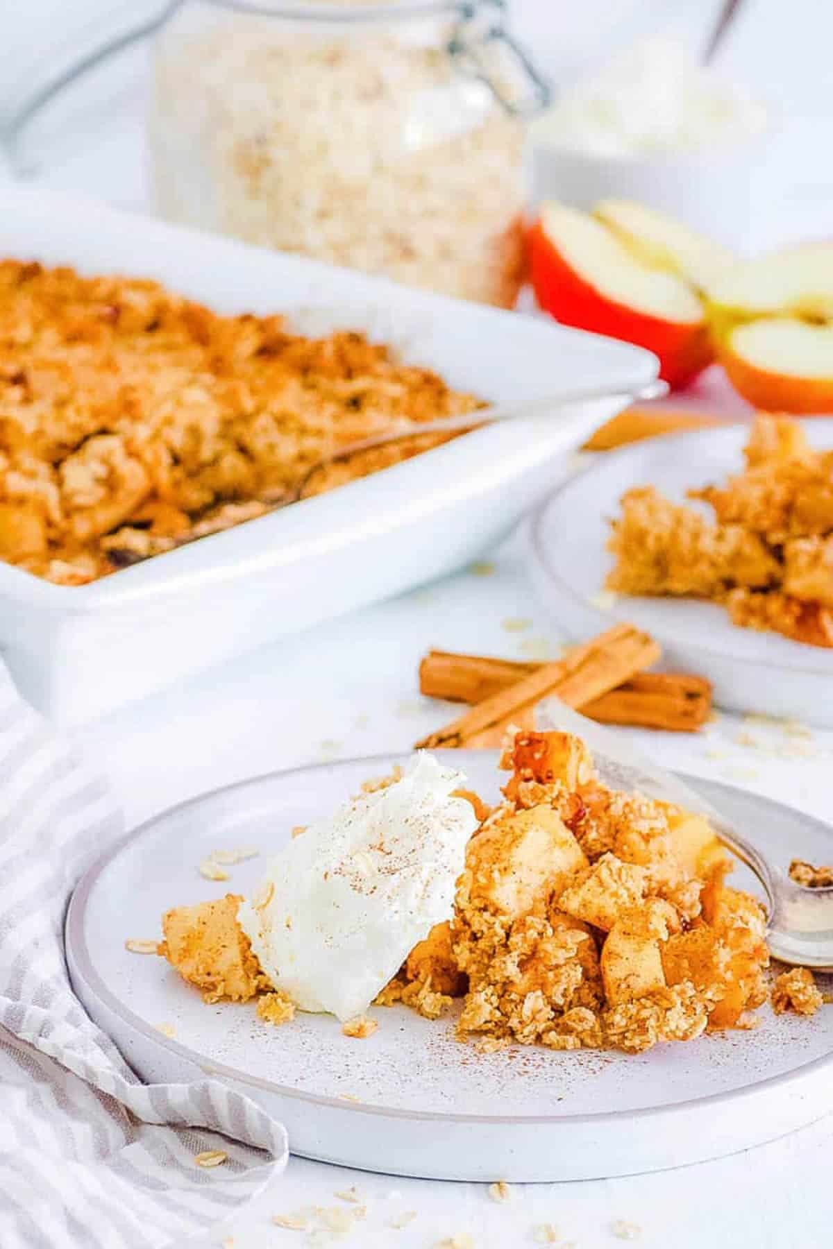 Side s،t of a plate of vegan apple crisp with a scoop of ice cream. A pan of the crisp sits in the background.