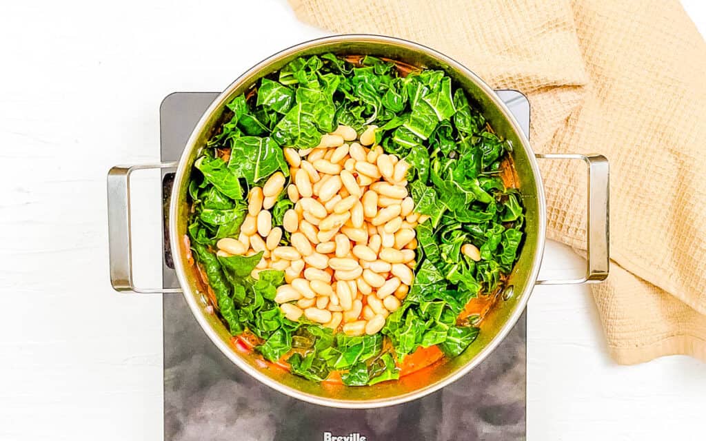 Cannellini beans and swiss chard added to soup ingredients in a pot on the stove.