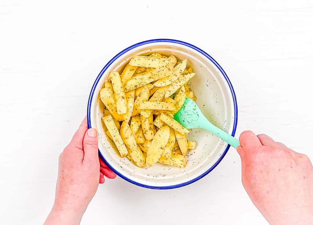 Potato wedges being tossed with Greek seasonings in a mixing bowl.