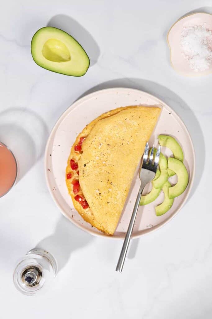 Vegan silken tofu omelette filled with tomatoes, on a pink plate with a silver fork and avocado slices.