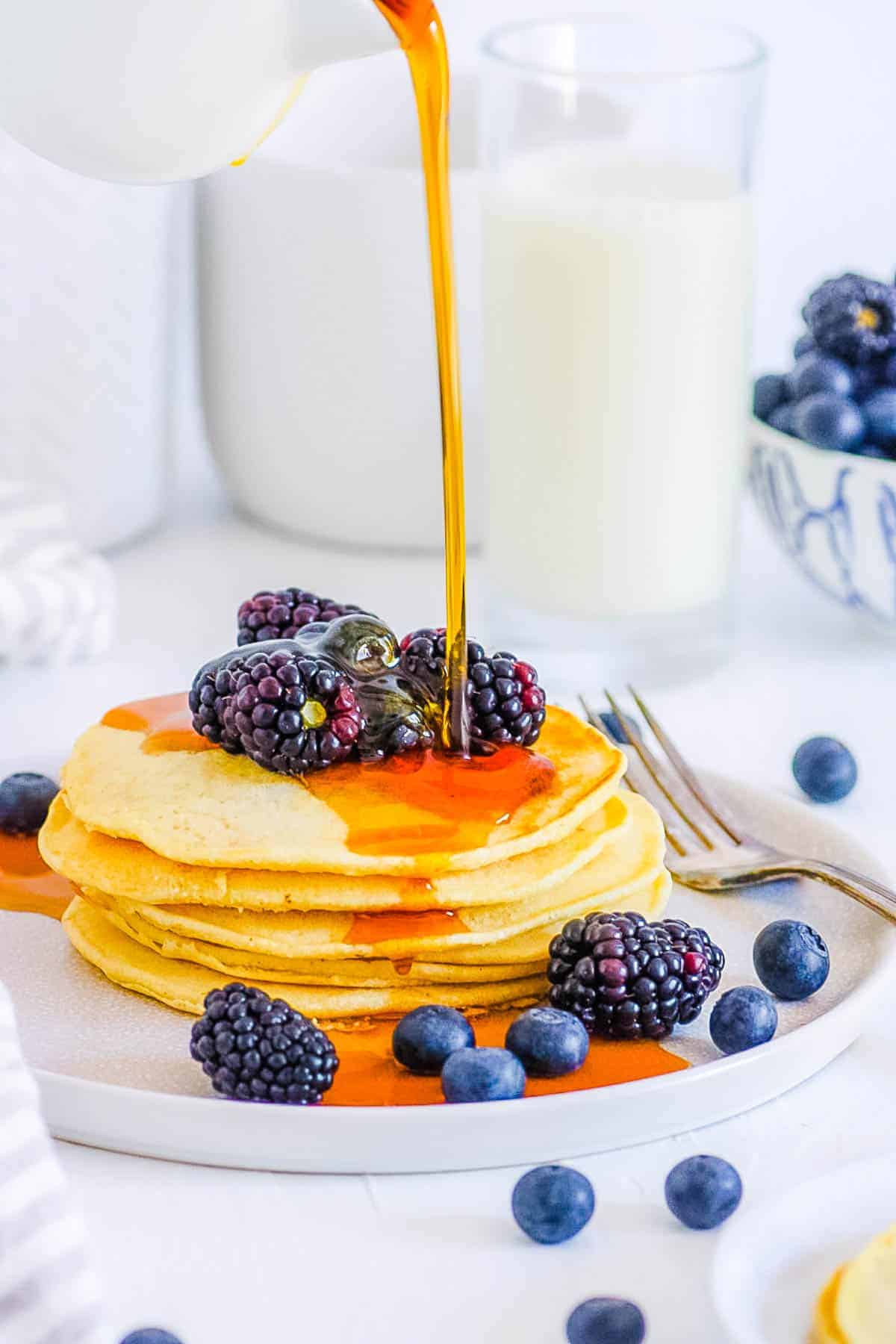 Maple syrup pouring onto coconut flour pancakes stacked on a white plate with blackberries and blueberries.
