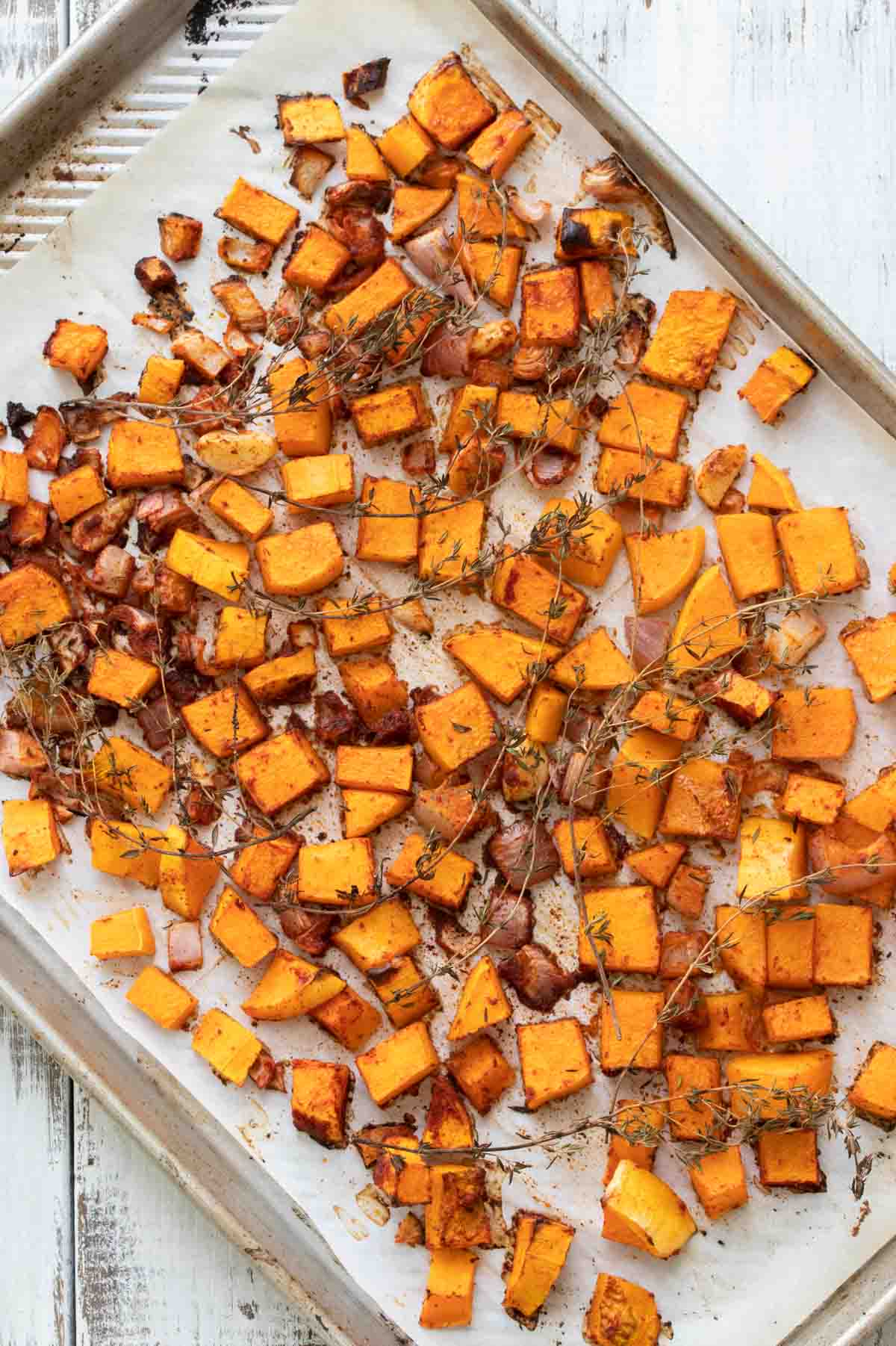 A baking pan with parchment and roasted pieces of butternut squash, thyme and shallots.