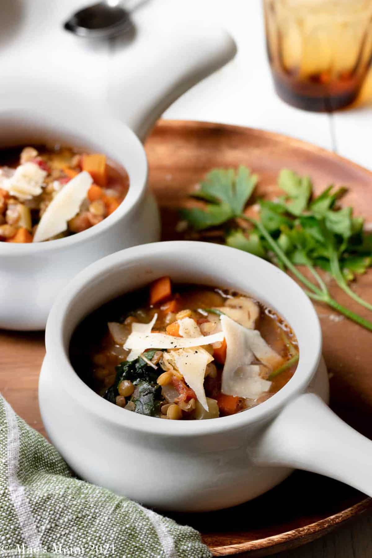 An angled shot of two cups of Italian lentil soup on a wooden cutting board with herbs.