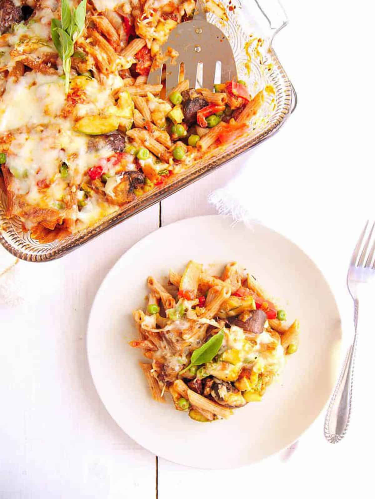 Baked vegan pasta with roasted vegetables, served on a white plate with a baking dish in the background.