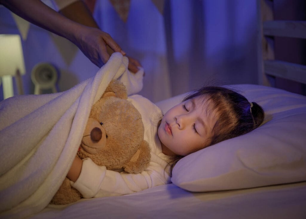 Mother putting blanket on and stroking head for her little girl daughter on bed in a dark bedroom at night.