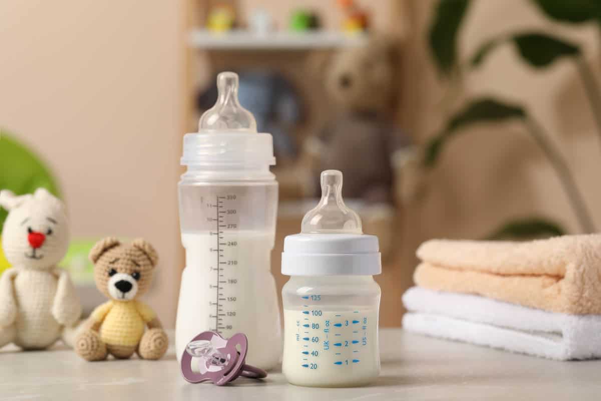 Feeding bottles with baby formula, pacifier, toys and towels on light grey table indoors.