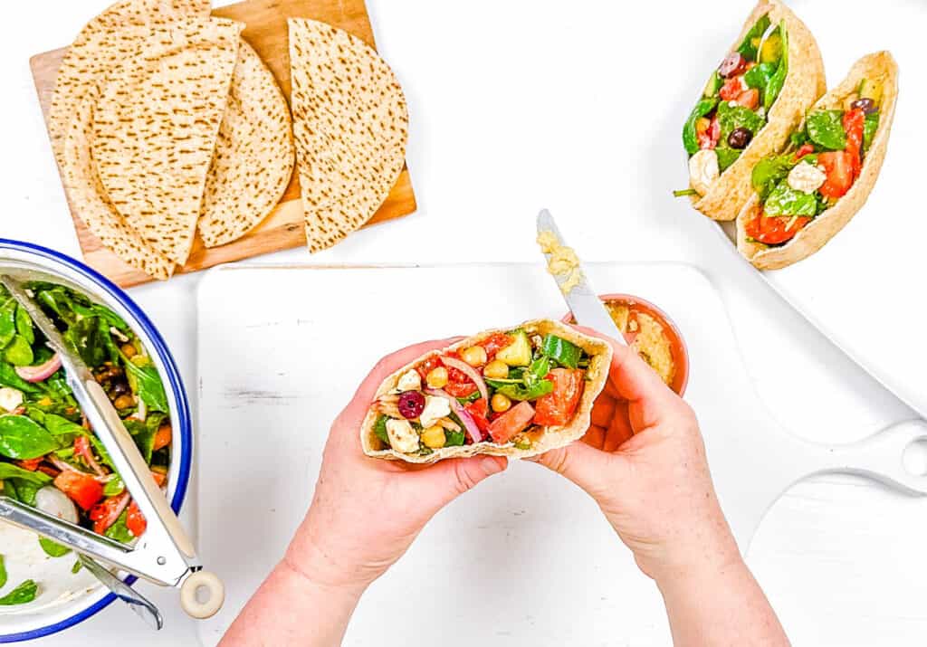 Greek pita sandwich stuffed with veggies, feta, and chickpeas on a white cutting board.
