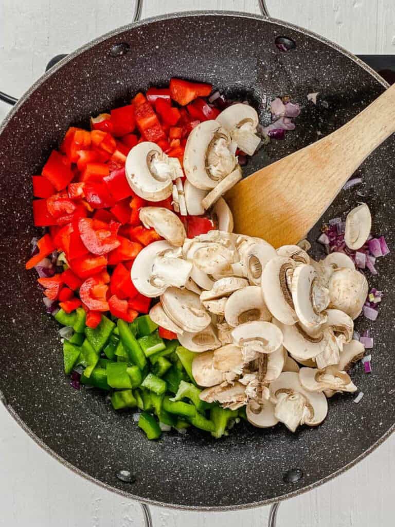 Cooking mushrooms, peppers, and onions in a wok with a wooden spoon.