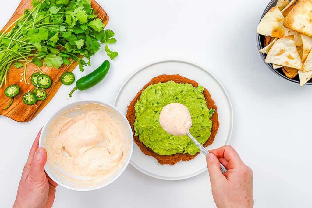 Greek yogurt added to layers of refried beans and guacamole on a white plate.