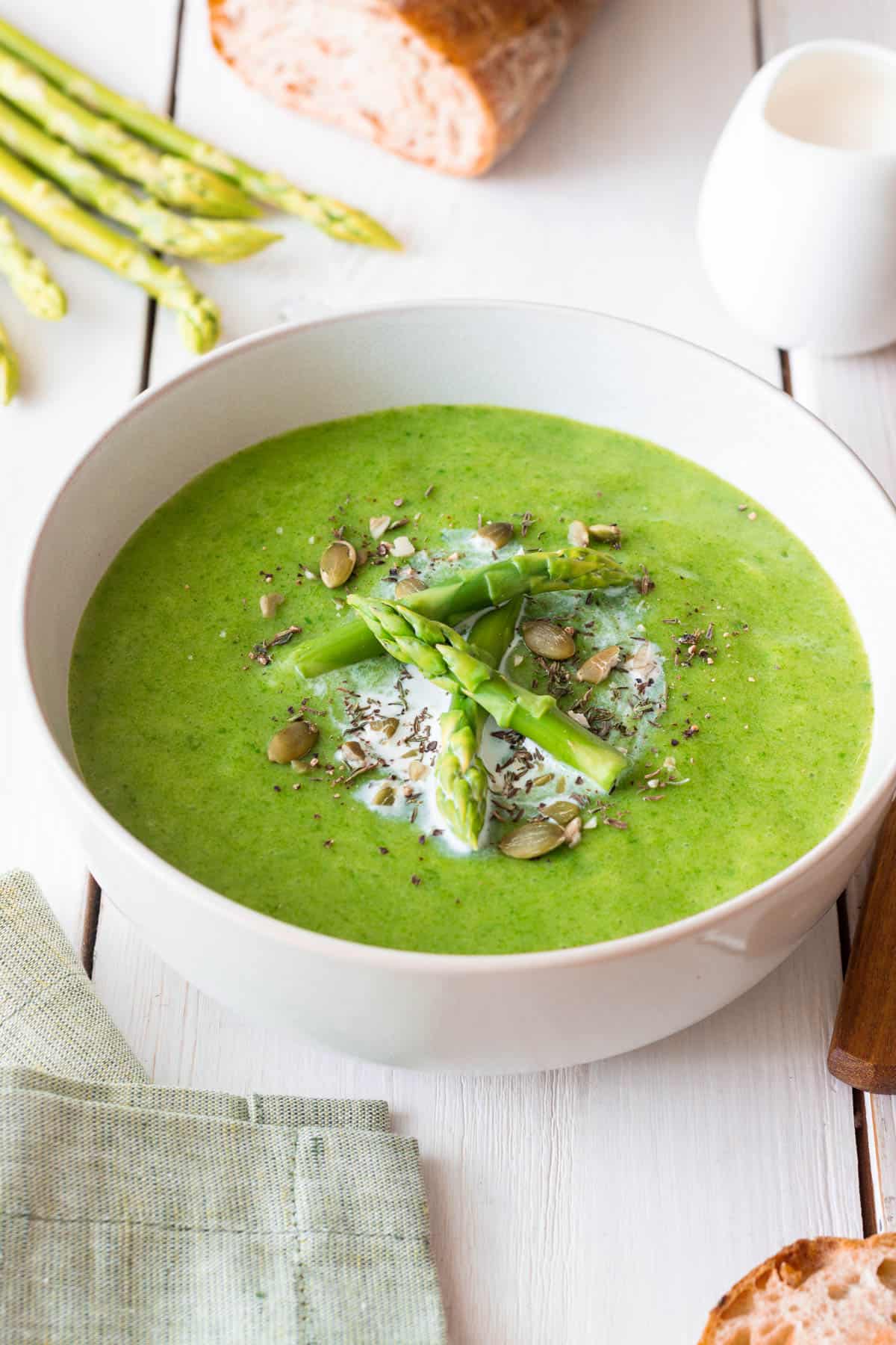 Easy asparagus soup without cream served in a white bowl, garnished with asparagus spears and pumpkin seeds on a white background.