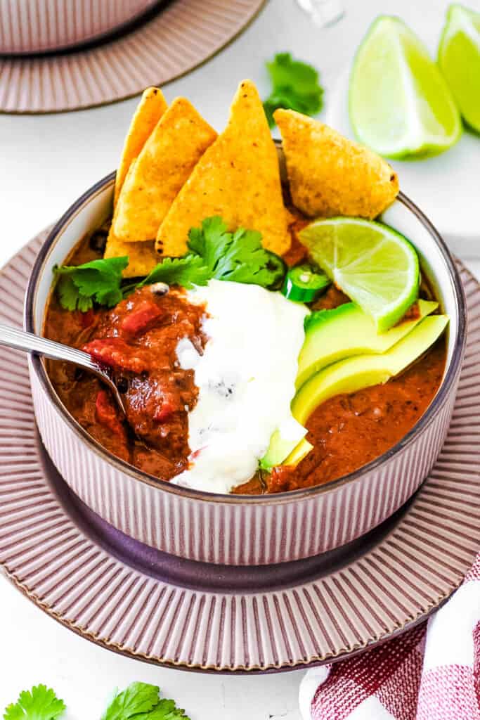 Easy vegan black bean soup, served in a grey bowl, garnished with cilantro, lime, avocado, crema, and corn chips.