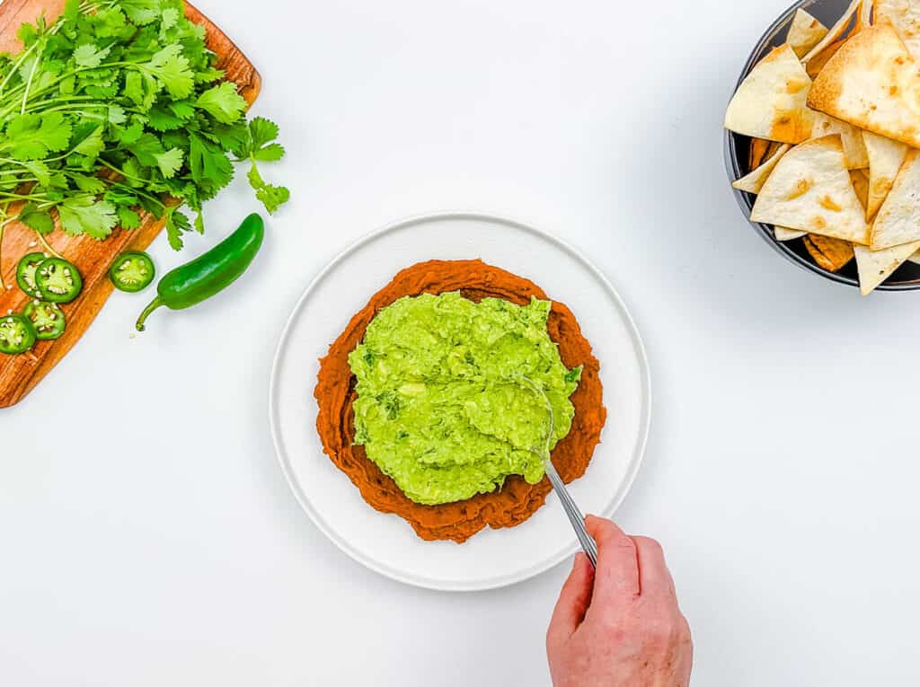 Guacamole layered onto refried beans on a white plate.