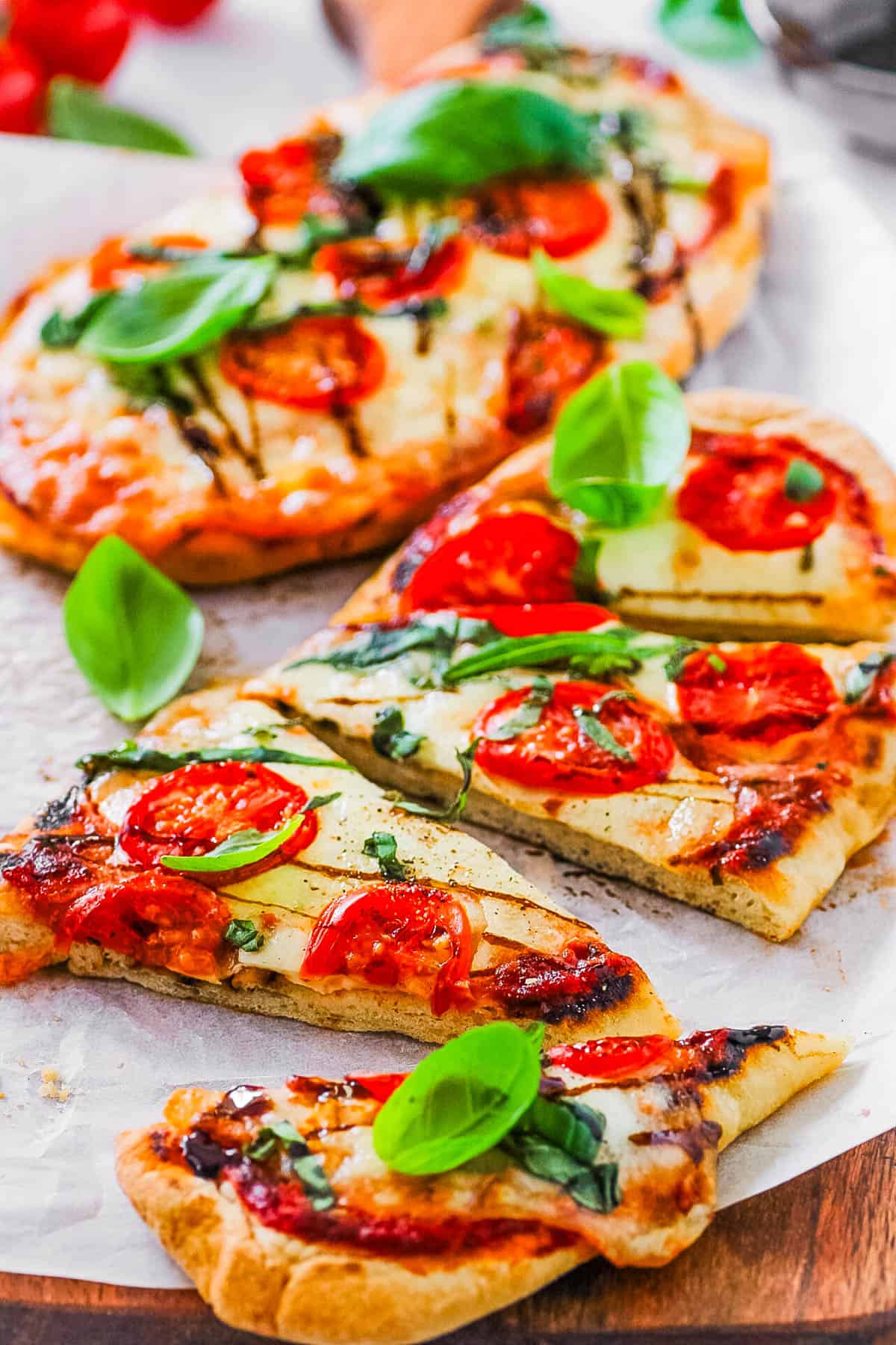 Margherita flatbread pizzas on a wooden cutting board, topped with basil, tomatoes and mozzarella.