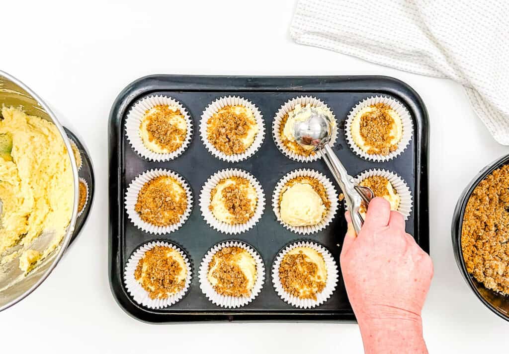 Batter for cinnamon muffins with a crumb topping being scooped into a muffin tin.