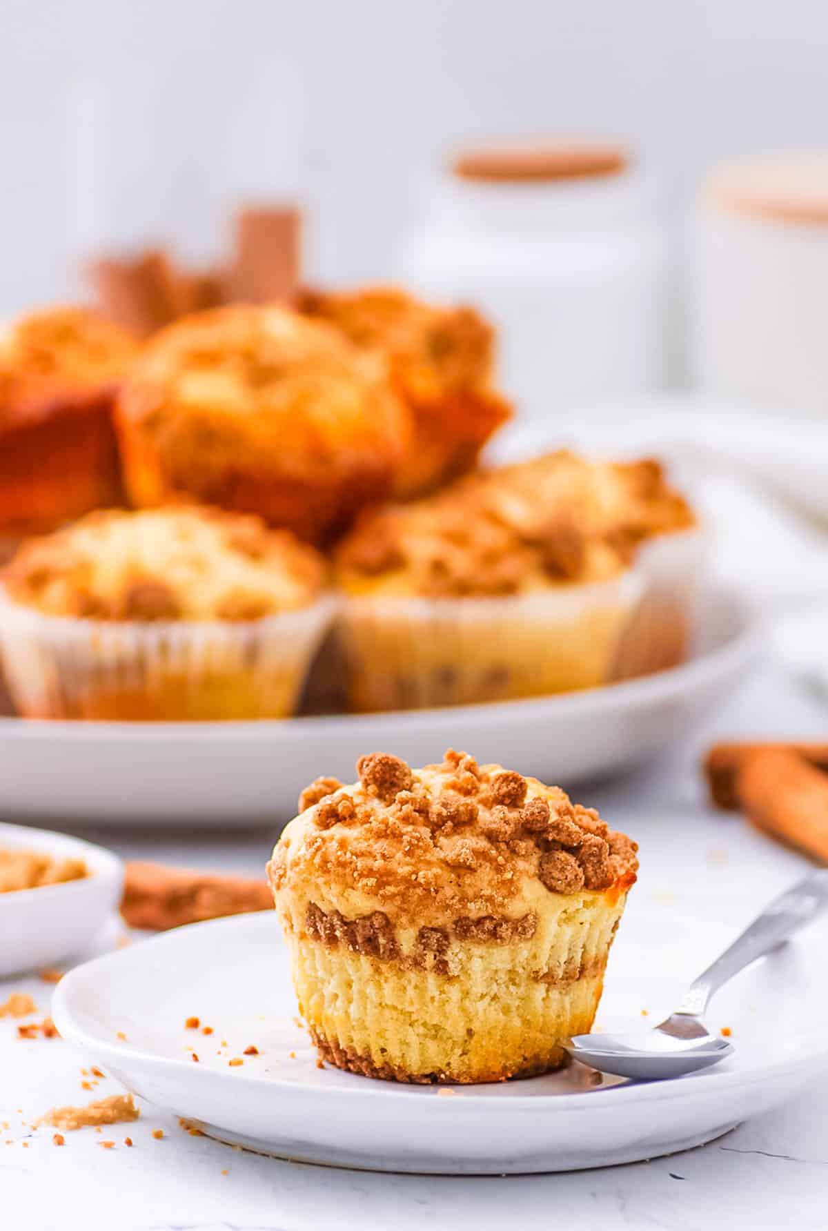 Cinnamon streusel muffin served on a white plate, with a bunch of baked cinnamon muffins with streusel topping in the background.
