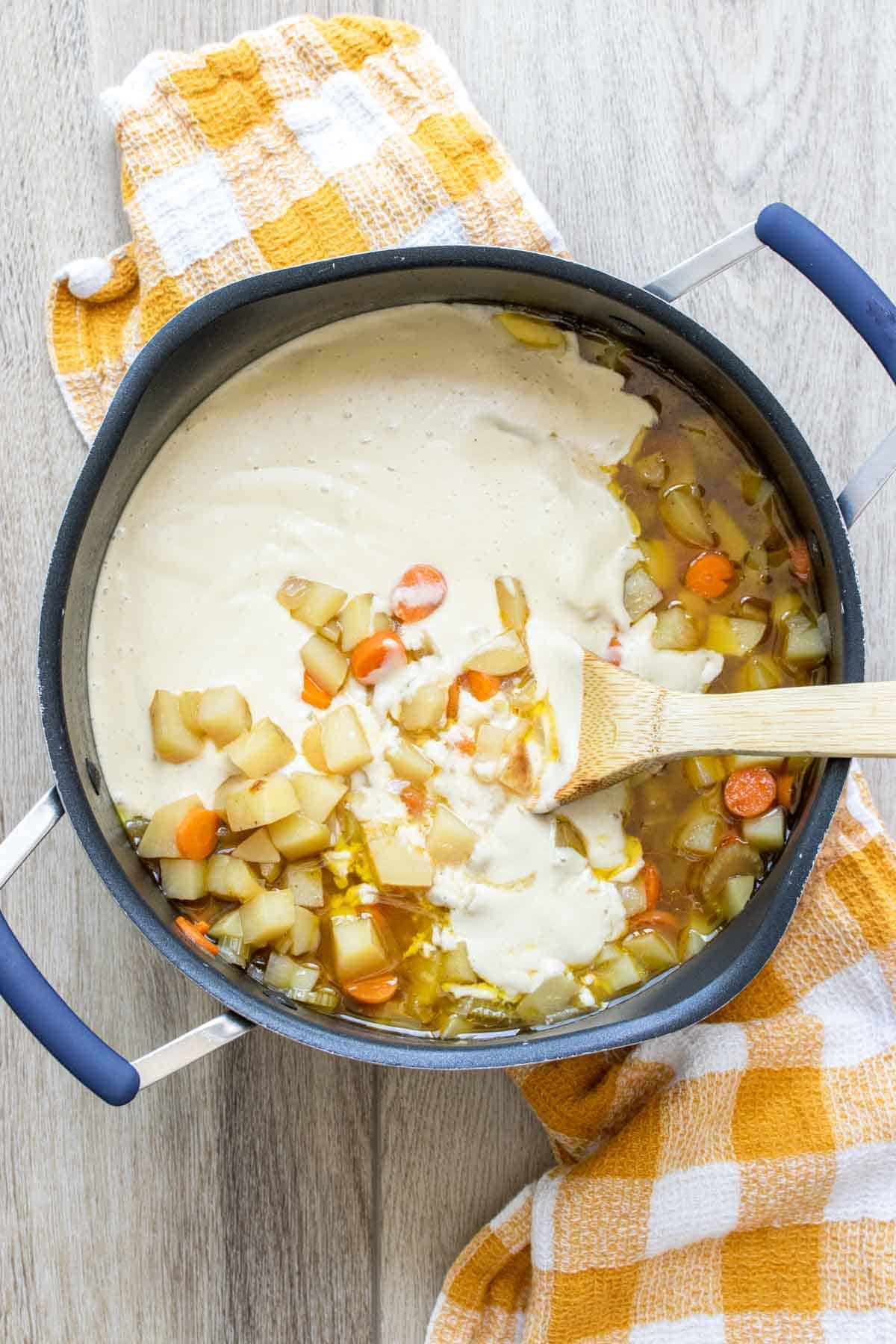 Wooden spoon mixing cream into a bowl of broth with veggies.