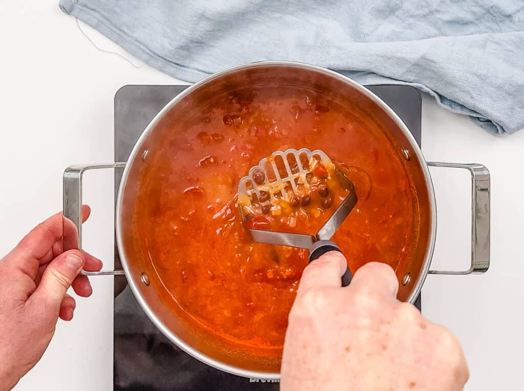 Hand with potato masher mashing black bean soup in a large pot on the stove.