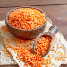A bowl of red lentils with a scoop on a towel-lined wooden counter.