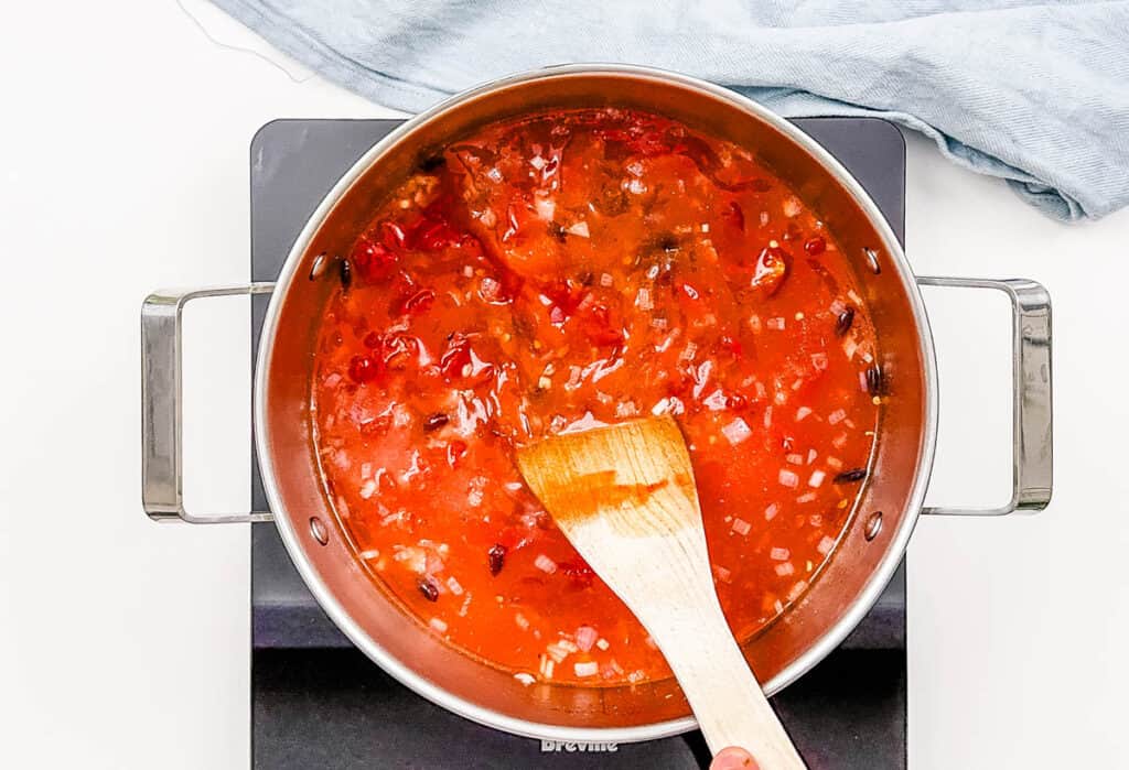 Tomatoes and broth added to black beans and veggies in a pot on the stove.