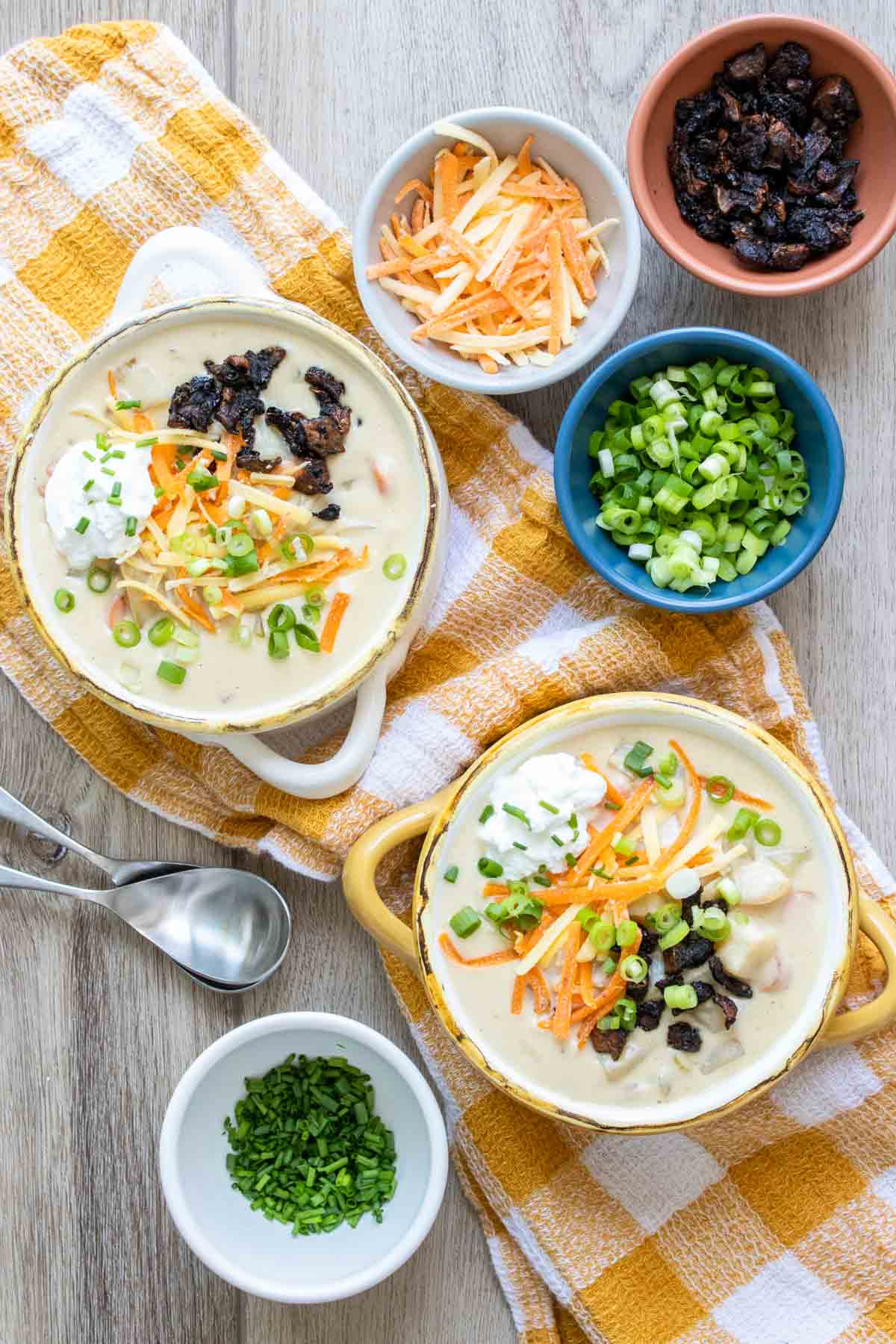 Two bowls of fully loaded vegan baked potato soup on a yellow and white checkered towel next to toppings.