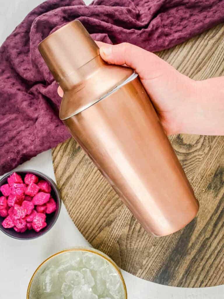 A hand shaking a rose gold colored cocktail shaker over a wooden cutting board, cups of fruit, and towel.