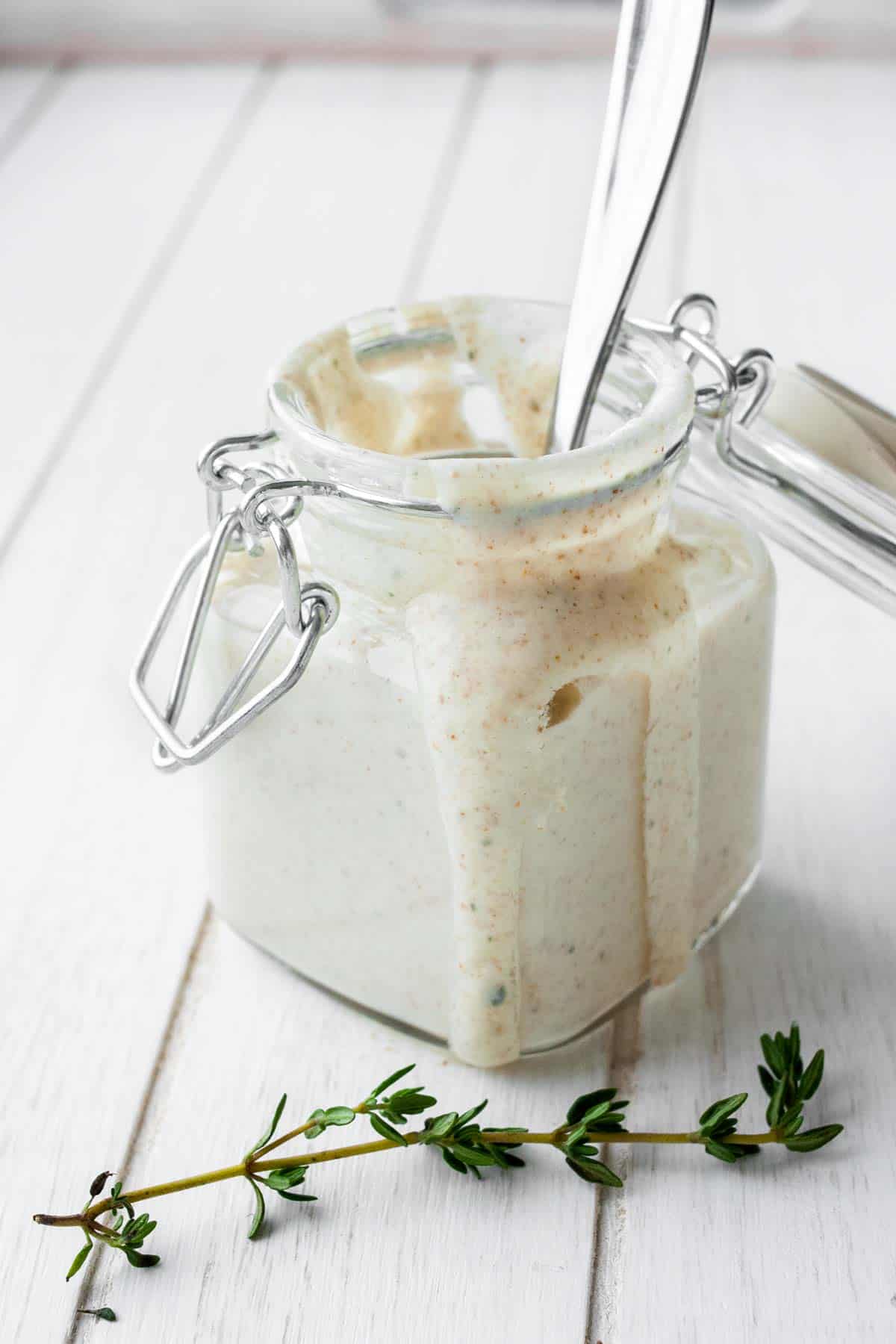 Front view of a thyme sprig in front of a glass jar of creamy sauce with a spoon in it.