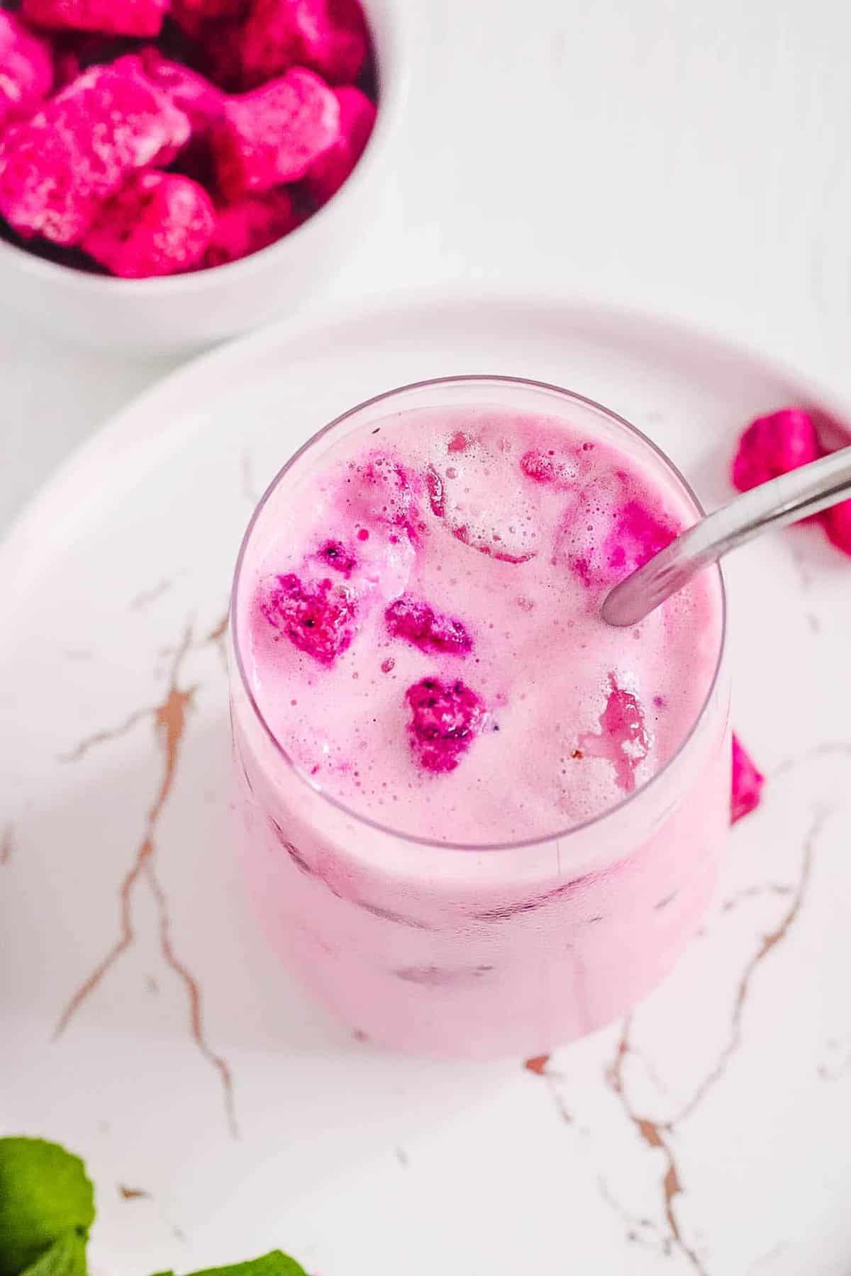 Overhead angled shot of a glass of dragon drink with a metal spoon next to a small dish of cubed dragon fruit.