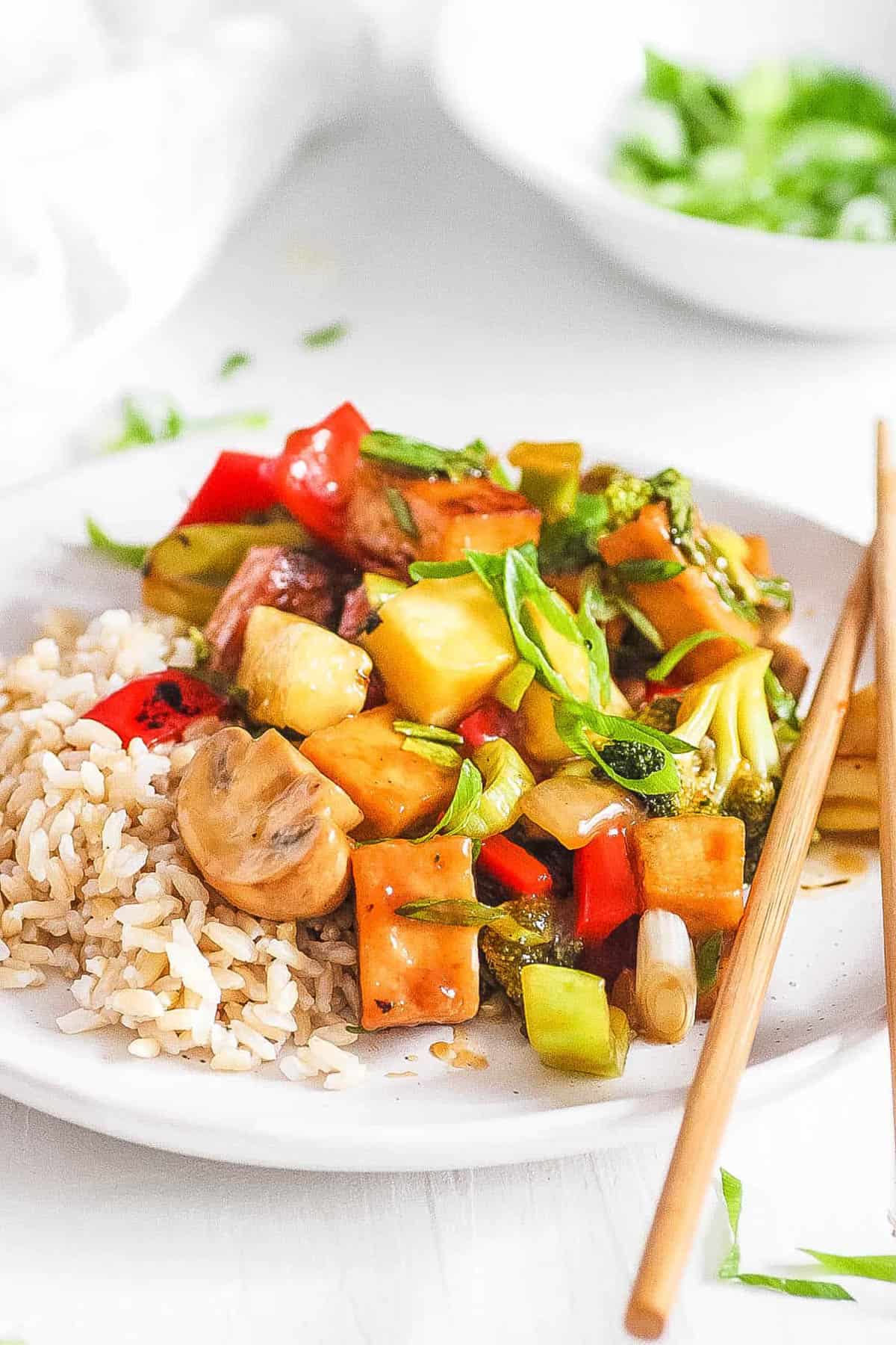 Sweet and sour tofu with veggies, served on a white plate with c،psticks.