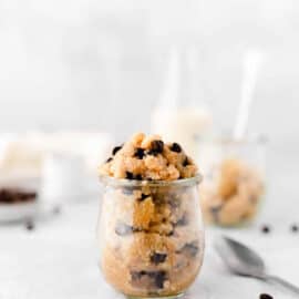 A side shot of a small cup of edible vegan cookie dough on the counter with chocolate chips, a spoon, and other cups of cookie dough.