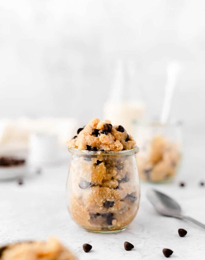 A side shot of a small cup of edible vegan cookie dough on the counter with chocolate chips, a spoon, and other cups of cookie dough.