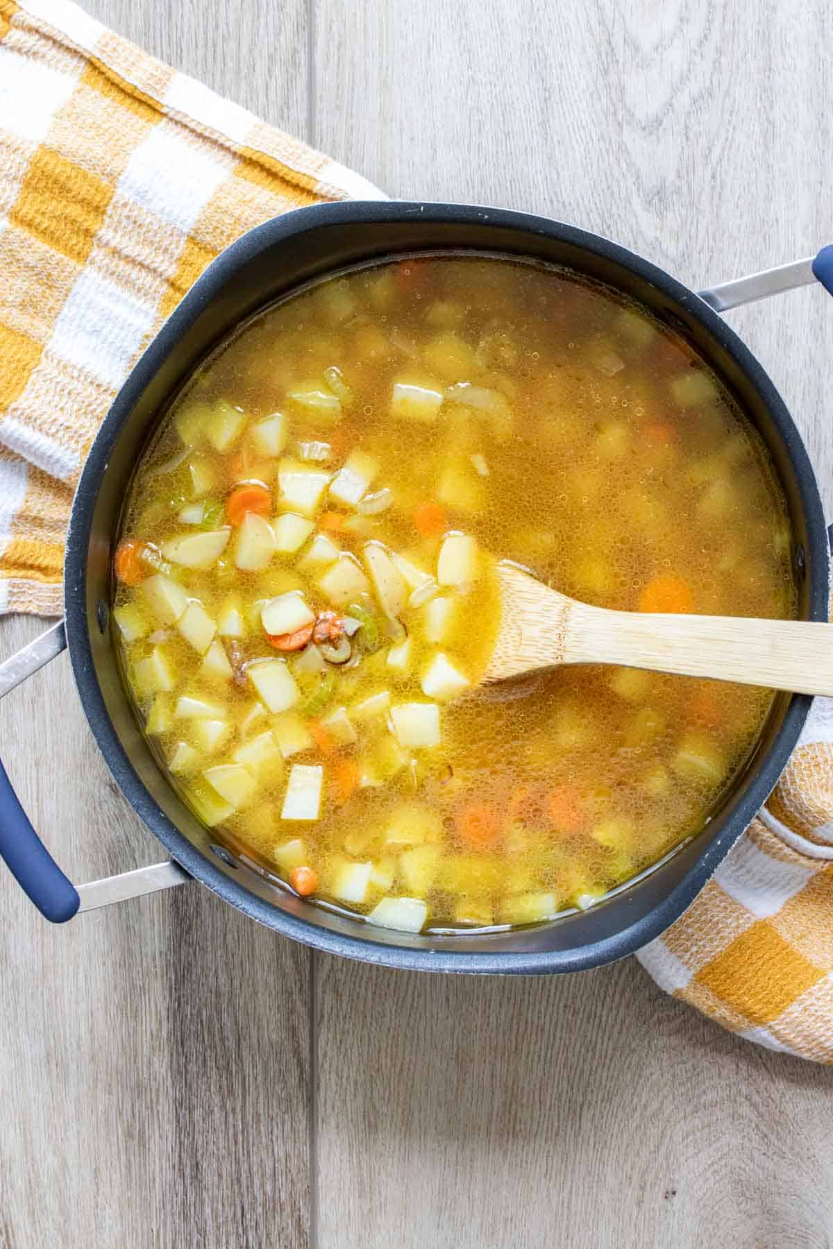 Pot filled with broth, chopped potatoes and carrots with a wooden spoon.