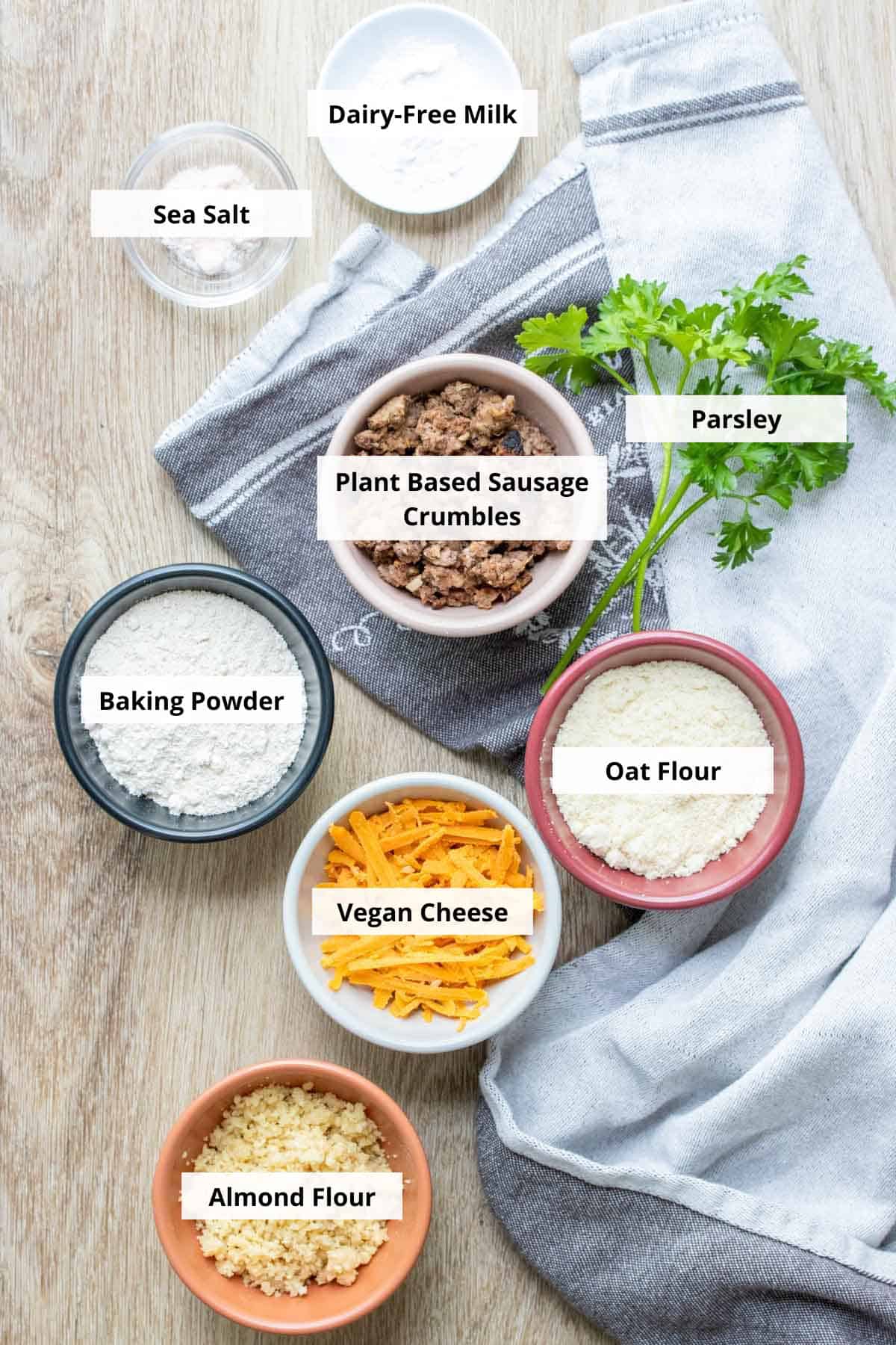 Ingredients for vegan sausage balls - bowls with flours, cheese, crumbled sausage baking powder and salt on a wooden surface with parsley.