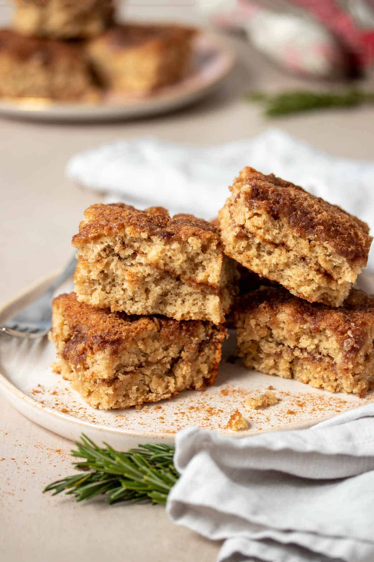 A tan plate with four pieces of coffee cake piled on it and speckled with cinnamon next to a white towel.